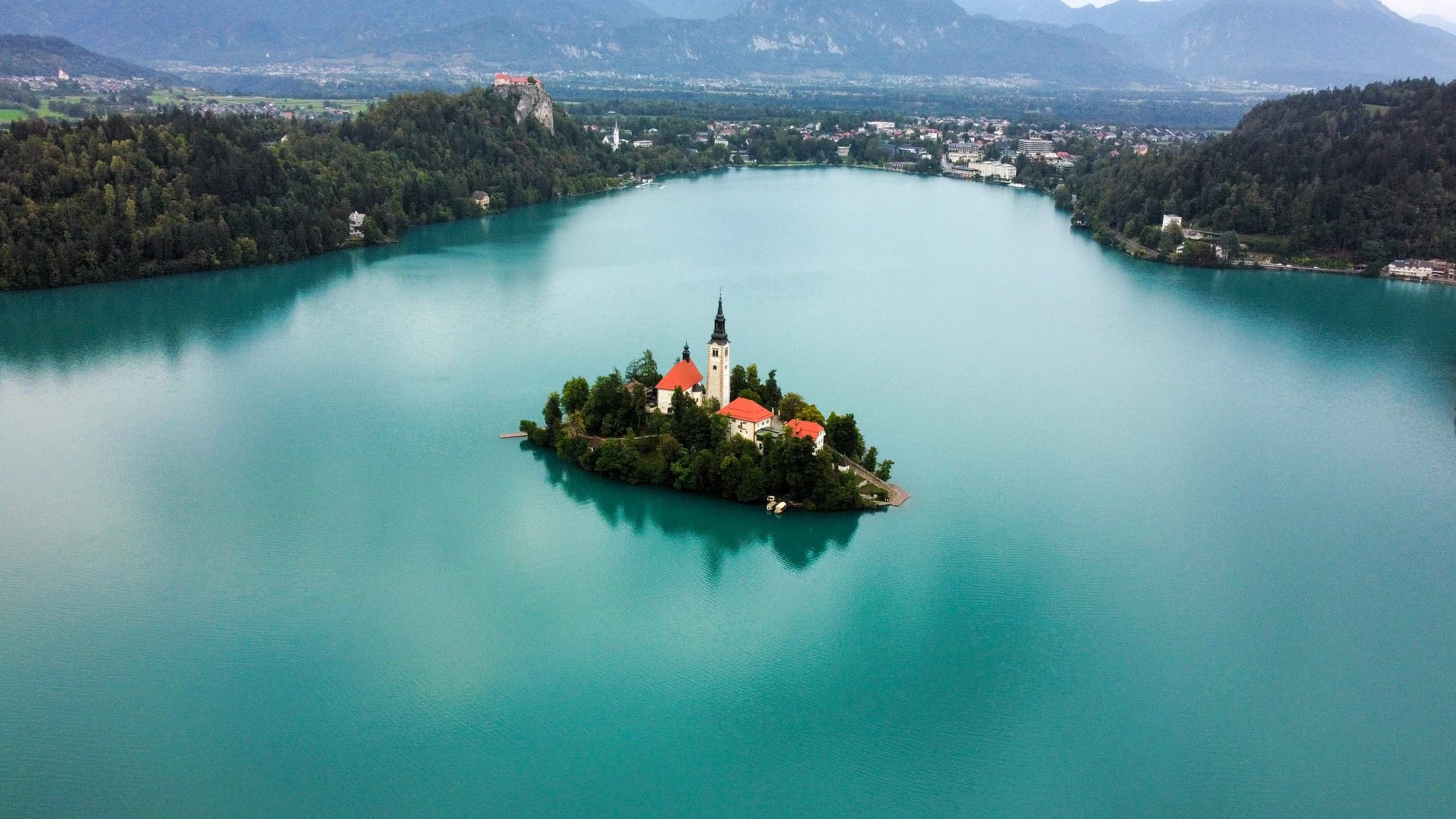 Aerial view of the church on the island in Lake Bled, surrounded by turquoise water and distant mountains.